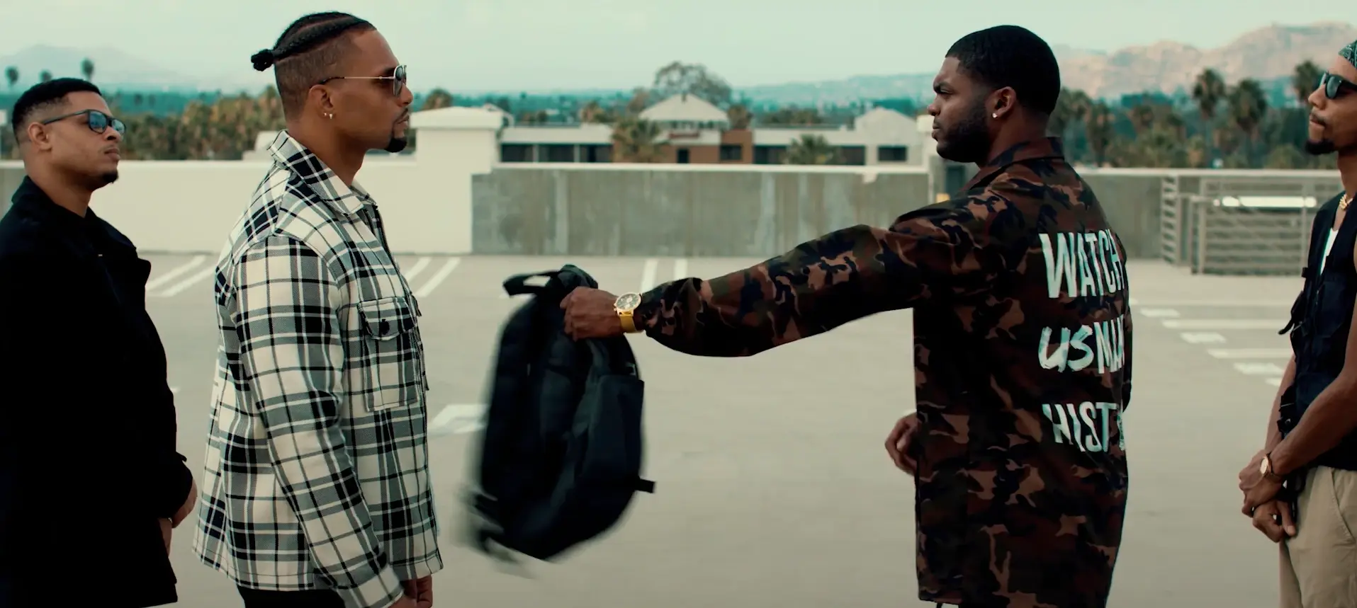 Four men face off on a rooftop parking structure; one hands over a black backpack.